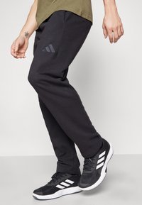 Juodos sportinės kelnės su tekstūruotu paviršiumi ir subtiliu tamsiai pilku Adidas logotipu ant šlaunies, derinamos su juodais sportiniais batais, turinčiais baltus akcentus.