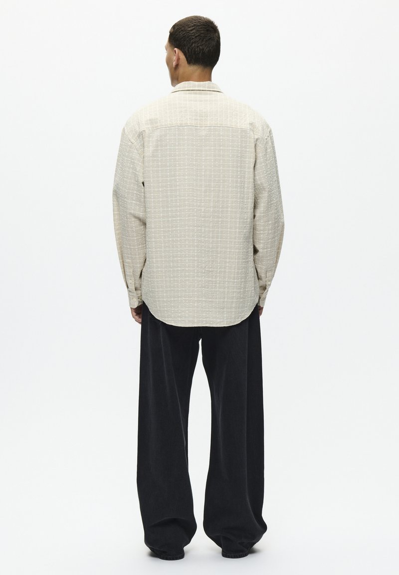Chemise longue à manches longues beige clair, texturée, avec un col à boutons, associée à un pantalon noir ample. Motif à carreaux simple sur la chemise.