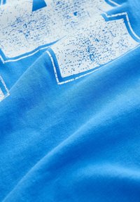 Sininen puuvillainen T-paita, jossa näkyy kankaan rakenne. Päälle painettu valkoinen pilkullinen graafinen kuvio sinisellä ääriviivalla.