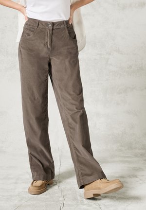 Pantaloni in velluto a coste marrone con design a gamba dritta, tasche frontali e chiusura a bottone. Indossati con scarpe texturizzate di un beige chiaro.