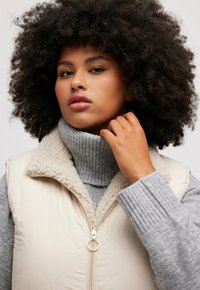 Cremfarbener gestepptes Weste mit einem beigen Fleecekragen, kombiniert mit einem grauen gerippten Rollkragenpullover. Das Model hat lockige Haare und eine Hand am Hals. Goldener Reißverschluss als Detail.