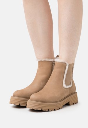 Bottines en cuir suédé marron clair avec une finition lisse, doublées de fourrure blanche sur le dessus, et une semelle épaisse rainurée pour une meilleure adhérence. Détail de fermeture éclair sur le côté.