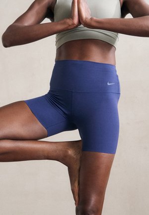 Persona con pantalones cortos Nike azules y sujetador deportivo verde claro de pie sobre una pierna en la postura del árbol de yoga con las manos unidas.