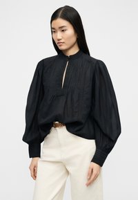 Γυναίκα με μακριά σκοτεινά μαλλιά που φοράει μαύρο πουκάμισο με μακριά μανίκια, ριγέ υφή και λαιμόκοψη με άνοιγμα, σε συνδυασμό με λευκό παντελόνι με ψηλή μέση και καφέ ζώνη.