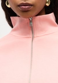 Γυναίκα που φοράει ανοιχτόροζ μπλούζα με φερμουάρ και ψηλό γιακά, με μεγάλα στρογγυλά χρυσά σκουλαρίκια, εμφανή τα χείλη και το κάτω μέρος του προσώπου.