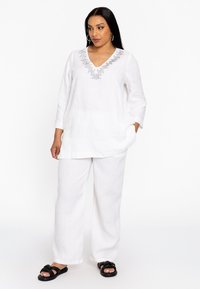 Witte linnen tuniek met een V-hals en decoratieve kraaldetails; gecombineerd met witte linnen broeken en zwarte sandalen.