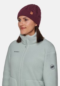 Bordowy dziany beanie z wzorem w warkocze oraz brązową skórzaną metką z logo, noszony z jasnoniebieską polarową kurtką z zapięciem na zamek.