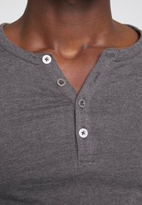 Graues kurzärmliges Henley-Shirt aus strukturiertem Stoff, mit einer Knopfleiste und drei silbernen Knöpfen sowie einem runden Halsausschnitt.