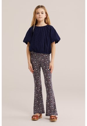 Mlada djevojka stoji noseći tamnoplavu bluzu s nabranim rukavima, cvjetno uzorkane široke hlače i smeđe sandale na platformu na jednobojnoj pozadini.