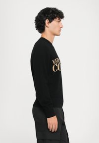 Melnā adīta džemperis ar ribotām aprocēm, uz kura priekšējās daļas ir izgaismoti burti "VERSACE" zeltainā krāsā, pieguļošs piegriezums un apaļš kakls.