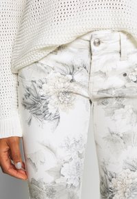 Białe dżinsy w kwiatowy wzór z teksturowanej tkaniny i kieszeniami. Charakteryzują się subtelnym szarym wzorem kwiatowym oraz srebrnymi akcentami.