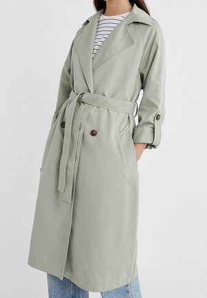 Femme portant un trench-coat vert clair ceinturé sur une chemise rayée et un jean bleu, mains dans les poches du manteau, sur fond uni.