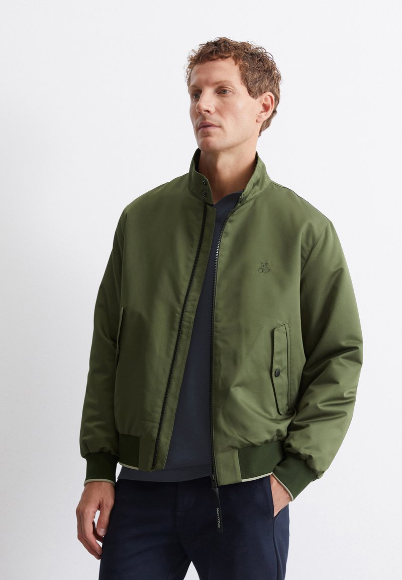 Olive green bomber jacket with a front zipper, standing collar, side pockets, ribbed cuffs, and a subtle logo on the chest.