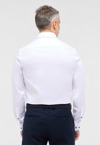 Witte blouse met lange mouwen en een puntige kraag, gladde textuur en contrasterende knopen, gedragen met marineblauwe broek en een gevlochten riem.
