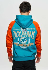Huvtröja med turkos kropp och oranga ärmar. Rygggrafik visar "Dolphins", Miami-logotypen och solstråle-design.