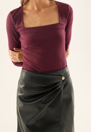 Femme portant un haut prune ajusté à manches longues avec un col carré et une jupe noire en cuir synthétique taille haute avec détail de boutons sur le côté.