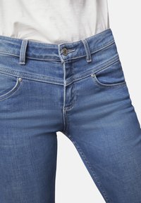 Los jeans de mezclilla azul cuentan con un corte ajustado, un diseño estándar de cinco bolsillos, y un cierre de botón y cremallera, con un ligero desgaste en la parte delantera.