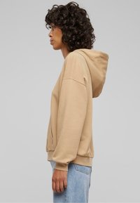 Sudadera beige con capucha ajustable, hombros caídos y puños acanalados. Combinada con jeans rectos de un azul claro. Textura suave.