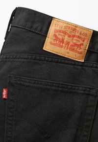 Jeans in denim nero con un'etichetta in pelle beige. L'etichetta presenta testo e immagini rossi, tra cui due cavalli. Logo Levi's rosso piccolo visibile.