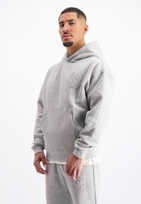 Grijze hoodie met een grote voorkantzak en geborduurd logo. Zacht katoenen materiaal, relaxte pasvorm en ribgebreide manchetten. Gecombineerd met bijpassende broeken.