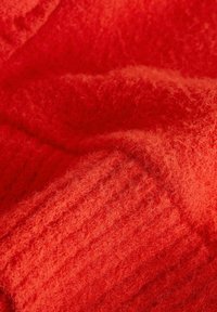 Rdeče pleteno blago z mehko, puhasto teksturo. Ima rebrast vzorec z različnimi debelinami, ki prikazuje globoko, živo barvo.