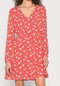 Punainen kukkamekko, jossa on pitkät hihat, V-pääntie, vyötärösolki ja kireä vyötärö. Sisältää pieniä valkoisia kukkia tekstuurilla taustalla.