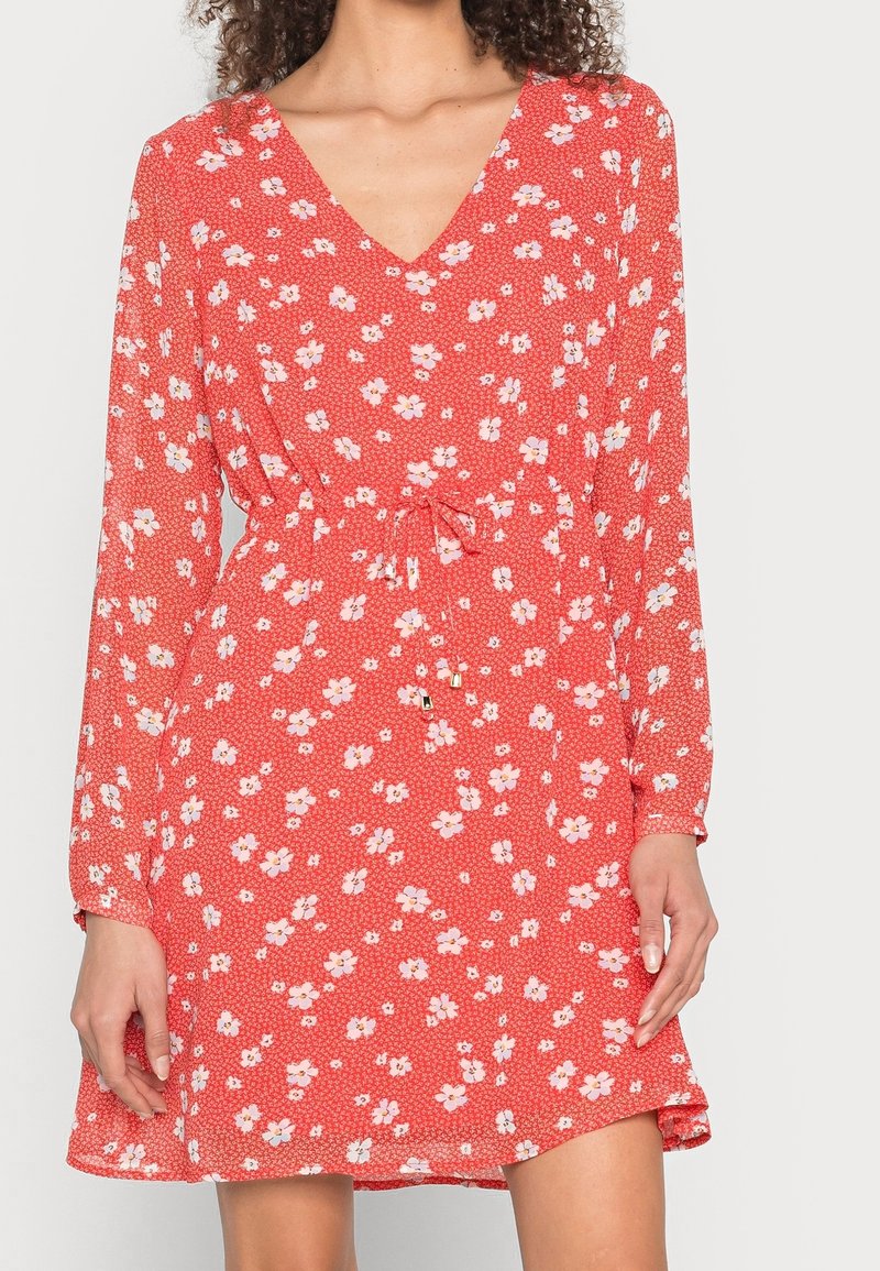 Punainen kukkamekko, jossa on pitkät hihat, V-pääntie, vyötärösolki ja kireä vyötärö. Sisältää pieniä valkoisia kukkia tekstuurilla taustalla.