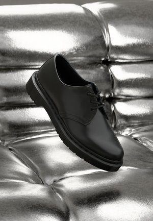 Chaussure en cuir noir avec un bout arrondi et des lacets, dotée d'une semelle en caoutchouc épaisse et texturée, reposant au-dessus d'une surface métallisée capitonnée.
