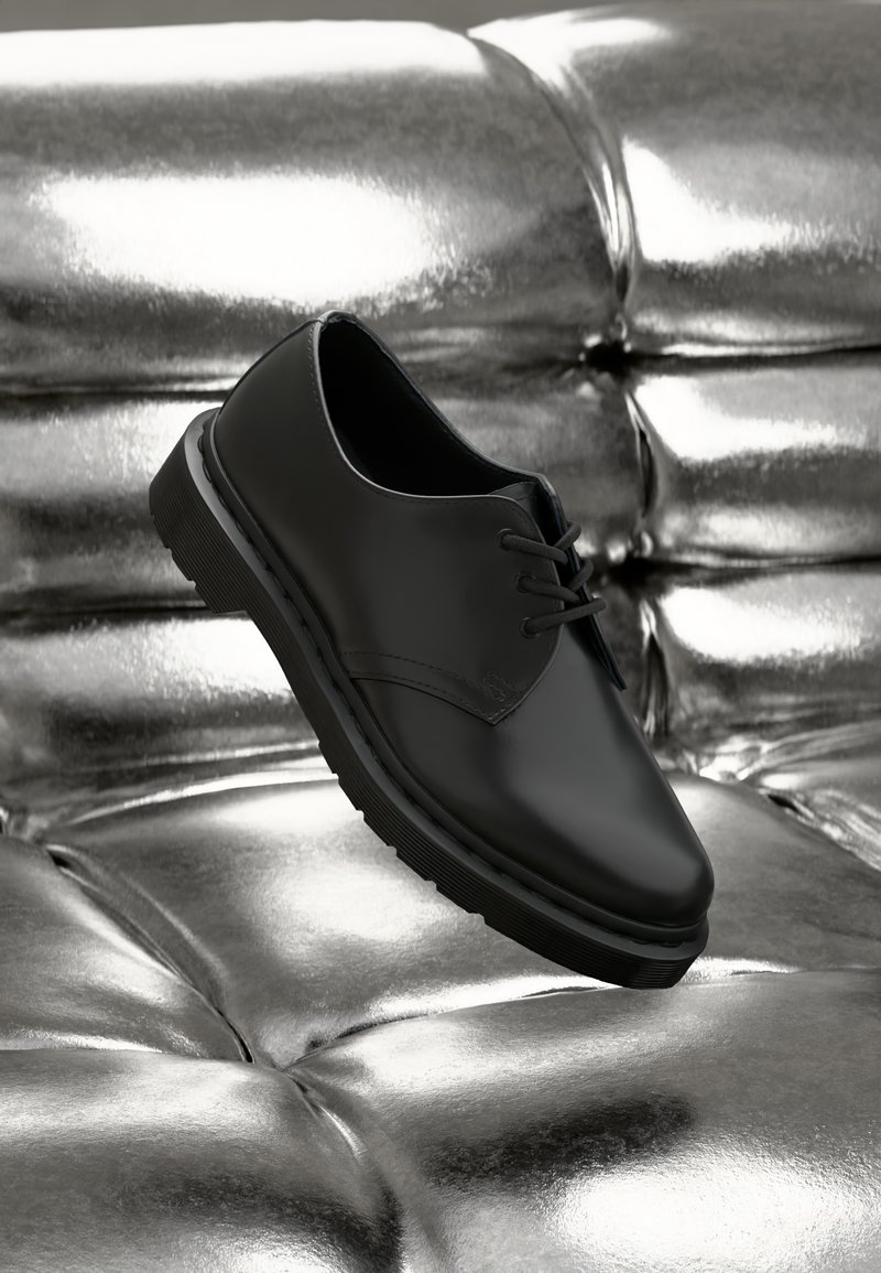 Black leather shoe with a rounded toe and laces, featuring a thick, textured rubber sole, resting above metallic tufted surface.
