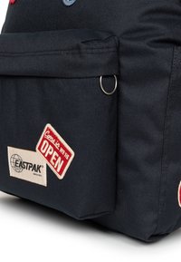 Mochila de tela negra con un bolsillo frontal, cierre de hardware plateado y parches con "Ven, ESTAMOS ABIERTOS" y el logo de Eastpak.