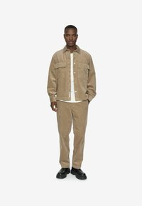 Beige corduroy jacket with two chest pockets, button closure, paired with matching pants and black footwear. Simple white shirt underneath.