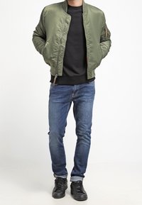 Chaqueta bomber color verde oliva, suéter de cuello redondo negro, jeans de mezclilla azul y zapatillas negras con superficie texturizada. La chaqueta tiene bolsillos con cremallera.