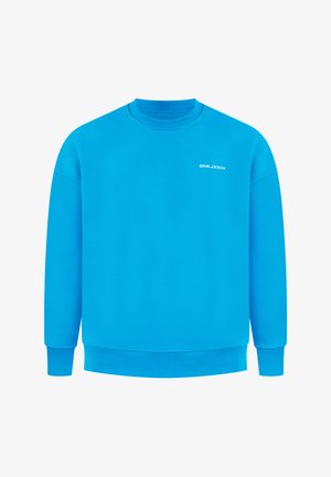 Mann trägt blauen Pullover mit weißem Motiv auf dem Rücken, schwarze Hose und weiße Sneakers, steht mit dem Rücken zur Kamera und schaut nach links.