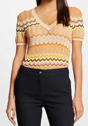 Frau trägt ein kurzärmeliges, gestricktes Top mit V-Ausschnitt in Orange, Gelb und Braun mit Zickzackmustern, kombiniert mit einer schwarzen Hose.