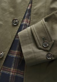 Manche de veste vert olive avec poignet boutonné sur tissu de chemise à carreaux bleu marine et marron.