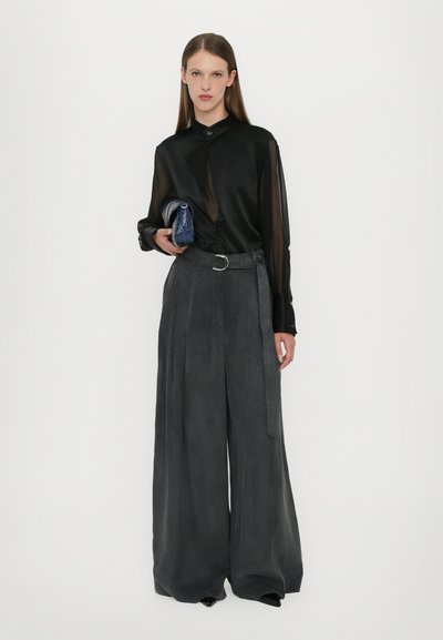 Blusa nera trasparente con colletto strutturato, abbinata a pantaloni grigi a gamba larga realizzati in tessuto morbido. Il modello tiene in mano una piccola pochette blu.