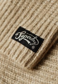 Beige strikket stof med ribbet tekstur, der har et sort mærke med ordet "Superdry" i sølvfarvet skrift.