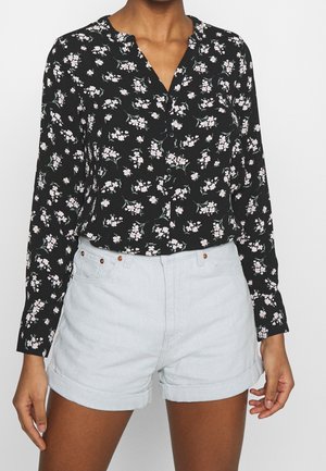 Femme portant un chemisier noir à manches longues avec un motif floral blanc et un short en jean bleu clair à ourlet roulé.