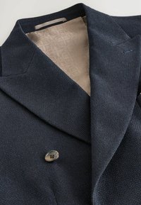 Námořnický blejzr s dvojitým zapínáním a texturovaným povrchem, s béžovou podšívkou s vzorem. Obsahuje široké klopy a jedno tlačítko na zapínání.
