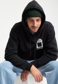 Sweat à capuche noir avec un design graphique sur la poitrine, porté avec un jean en denim bleu clair. Le modèle porte un bonnet vert.