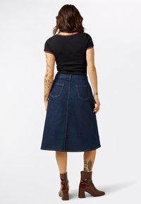 Tumman denim A-linjainen hame korkean vyötärön kanssa, takataskut ja ommeltuja yksityiskohtia, yhdistettynä tiukkaan mustaan t-paitaan ja ruskeisiin nilkkureihin.