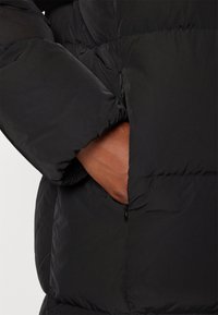 Black puffer jacket with quilted texture, featuring a zipper pocket on the side and a partially visible hand tucked into the garment.