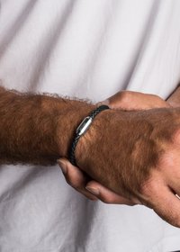 Fekete, fonott bőr karkötő ezüst fémcsattal, amelyet a csuklón viselnek. A dizájn texturált felületet és karcsú sziluettet mutat.