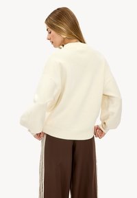Pull en crème surdimensionné avec un tricot texturé, un ourlet à côtes et des coutures d'épaules tombantes. Associé à un pantalon marron foncé avec des rayures blanches.