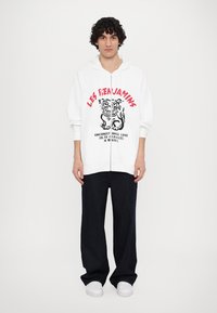 Λευκή φούτερ ζακέτα με σχέδιο και κείμενο, συνδυασμένη με χαλαρά σκούρα τζιν και λευκά αθλητικά παπούτσια.