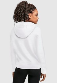Fehér kapucnis pulóver puha textúrával, kenguruzsebb és bordázott mandzsetták jellemzik. A modellt hátulról látjuk, fekete leggings-szel párosítva.