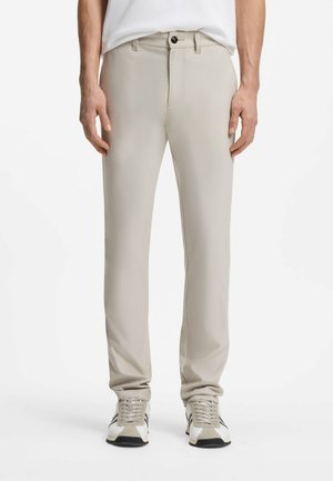 Mann trägt schlanke beiger Hose und weiße Sneakers mit beigen und schwarzen Details, steht vor einem einfarbigen Hintergrund.