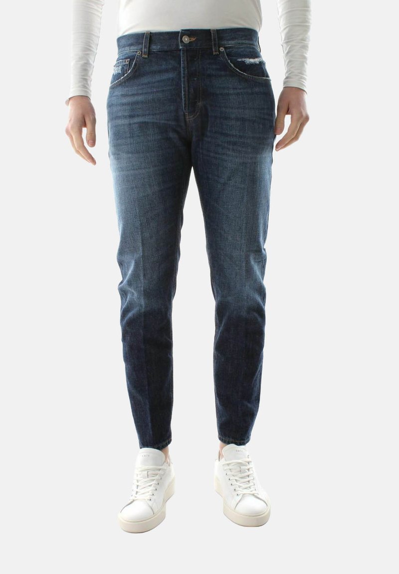 Jeans in denim scuro con vestibilità slim, aree sbiadite, tasche anteriori e chiusura con bottone, abbinati a sneakers bianche.