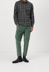 Geruit overhemd met knopen in donkere tinten, gecombineerd met groene corduroy broek. Model draagt zwarte enkellaarsjes. Strakke lijnen en gestructureerde pasvorm.