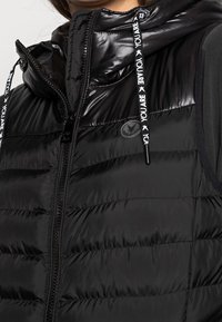 Czarna pikowana kamizelka z błyszczącym wykończeniem, z wysokim kołnierzem, zamkiem błyskawicznym i czarnymi sznurkami z białym napisem. Na piersi naszywka z logo.